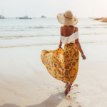Γυναίκα που περπατά στην παραλία με ψάθινο καπέλο και κίτρινο αέρινο φόρεμα με φλοράλ μοτίβο και λευκό τοπ που μοιάζει με μπλούζα - μοντέρνα φορέματα για το καλοκαίρι του 2023