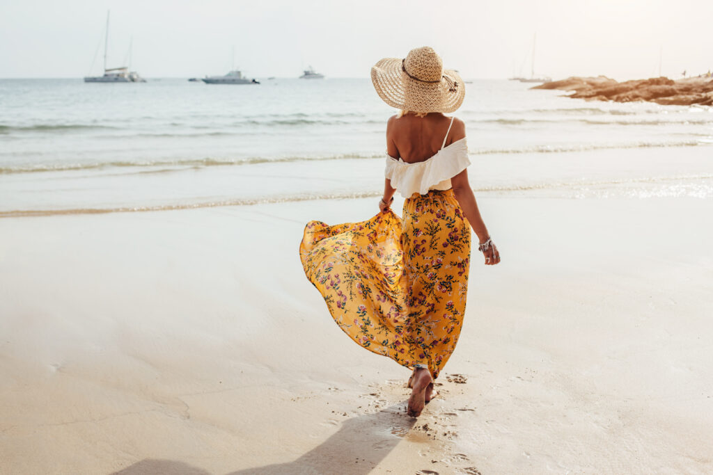 Γυναίκα που περπατά στην παραλία με ψάθινο καπέλο και κίτρινο αέρινο φόρεμα με φλοράλ μοτίβο και λευκό τοπ που μοιάζει με μπλούζα - μοντέρνα φορέματα για το καλοκαίρι του 2023